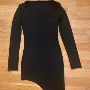 Svart asymmetrisk långärmad klänning med slits i storlek petite XS - Snygg svart klänning med lång ärm och asymmetrisk nederkant. Modellen är tight och har en fyrkantig ringning som ger en modern vibe. Materialet känns mjukt och stretchigt, perfekt för dig som gillar stilrena plagg med en twist.