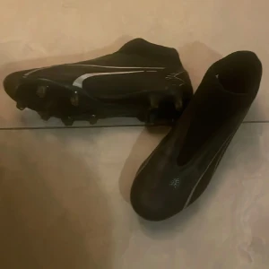 Svarta Puma fotbollsskor utan snörning str 44,5 - Säljer ett par svarta Puma fotbollsskor med vita swoosh-detaljer på sidorna. Skorna har en strömlinjeformad design utan snörning och är tillverkade i syntetmaterial för bra passform och grepp på planen. Perfekta för dig som vill ha stilrena och snabba skor till matchen. Sparsamt använda , storlek 44,5. 