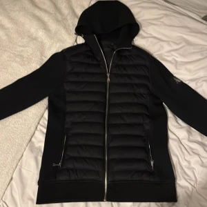 AZ Cardigan  - Svart cardigan från Alessandro Zavetti med huva och snygga quiltade paneler framtill och på axlarna. Jackan har silverfärgad dragkedja, två fickor med dragkedja och en stilren logotyp på ärmen. Perfekt för dig som vill ha en clean look.