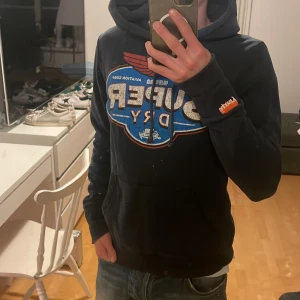 2000s Superdry hoodie (unikt tryck) - Svart hoodie från Superdry i storlek small med stort färgglatt tryck på bröstet i blått, rött och beige. Tröjan har huva med snörning, ribbade muddar och en klassisk känguruficka. Detaljerad Superdry-logga på ärmen och insidan. Perfekt för dig som gillar streetwear. Defekter förekommer. Finns fläckar från målarfärg därav ett överkomligt pris. Om färgfläckarna tillägger en vintage-känsla ligger i betraktarens ögon 😁🙌 Skriv vid funderingar 📲