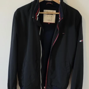 Svart bomberjacka från Tommy Jeans - Svart bomberjacka från Tommy Jeans med klassiska ribbade muddar i marinblått, rött och vitt. Jackan har dragkedja framtill, diskret logga på bröstet och ärmen samt sportig krage med röd och vit detalj. Tillverkad i polyester, perfekt för en stilren och sportig look.