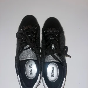 Svarta och vita sneakers Michael Kors - Snygga sneakers från Michael Kors med svart och vit färg, MK-logga på sidan och plös, samt vit platt sula. Skorna har snörning och ett diskret mönster med MK över hela ovandelen. Materialet är en mix av syntet och textil med silvriga detaljer vid hälen. MK märket på sko snöret går att plocka bort. Helt oanvända köpte en för stor storlek! Väldigt fint skick 