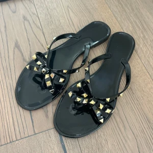 Svarta sandaler med nitar och rosett - Svarta sandaler med platt sula och öppen tå, dekorerade med en stor rosett framtill och guldiga pyramidformade nitar. Liknar känd sandal, men köpt från en skobutik i Italien. Så snygga men säljs då jag har för många skor. Oanvända.