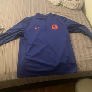 Blå långärmad fotbollströja Nike Storm-FIT - Säljer en blå långärmad fotbollströja från Nike med Storm-FIT-material. Tröjan har Nederländernas landslagsmärke och orange Nike-logga på bröstet. Perfekt för träning eller match. Tillverkad i Vietnam.