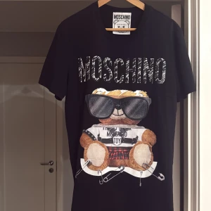 Svart Moschino t-shirt med nalletryck - Svart t-shirt från Moschino med stort tryck av en nallebjörn i solglasögon och cool outfit framtill. T-shirten har korta ärmar, rund hals och är gjord i mjuk bomull. Perfekt statement-plagg för dig som gillar streetstyle och designer.
