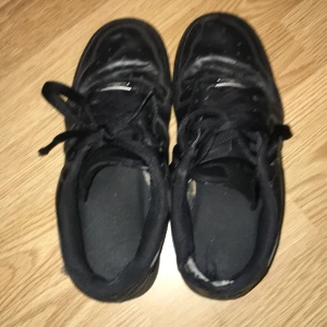 Svarta Nike Air Force 1 sneakers - Svarta Nike Air Force 1 sneakers med klassisk låg siluett och snörning. Skorna är tillverkade i skinn med platt sula och rund tå. Swoosh-loggan syns på sidan och designen är helt svart, vilket ger en clean och stilren look.