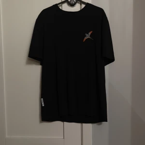 Svart t-shirt med fågeltryck - Svart t-shirt med kort ärm och en Axel Arigato fågel på bröstet. Klassisk rund halsringning och relaxed fit. Liten vit etikett nertill på sidan. Perfekt för en clean och stilren look.
