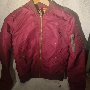 Vinröd bomberjacka från Schott - Säljer en vinröd bomberjacka från Schott med orange foder och klassisk guldfärgad dragkedja. Jackan har ribbade muddar, fickor med tryckknappar och en ärmficka med dragkedja. Märkespatch på ena ärmen och snygg, sportig look.