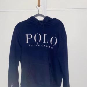Mörkblå hoodie från Ralph Lauren - Säljer en mörkblå hoodie från Ralph Lauren med stor vit POLO-logga framtill. Tröjan har huva med snörning och en klassisk känguruficka. Perfekt för dig som gillar stilrena och bekväma plagg med premiumkänsla. PRIS KAN DISKUTERAS!