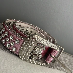 Glittrigt rosa bälte med döskalle - Säljer ett rosa bälte med glittrig yta och stora silvriga nitar samt strassdetaljer. Spännet är dekorerat med stenar och har flera coola döskallar. 100cm. Vet tyvärr inte vart det kommer ifrån då jag fick det för länge sen. Använt runt 1-2 gånger och funkar perfekt. 