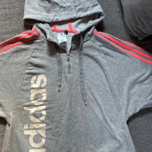Grå hoodie från Adidas med rosa detaljer - Snygg grå hoodie från Adidas med rosa ränder längs ärmarna och stor logga på framsidan. Tröjan har huva, halv dragkedja och är tillverkad i mjuk bomullsmix. Perfekt för en sportig och avslappnad stil.
