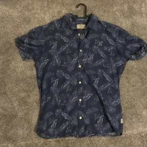 Blå mönstrad skjorta Jack & Jones - Snygg kortärmad skjorta från Jack & Jones i marinblått med tropiskt bladmönster. Skjortan har klassisk krage, knappar framtill och är tillverkad i mjuk bomull. Perfekt för dig som gillar chill och somriga vibes.