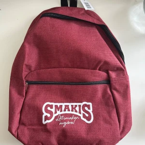 Röd ryggsäck - oanvänd - Snygg bordeauxfärgad ryggsäck från Smakis med vit logga framtill. Väskan har ett stort huvudfack, en framficka med dragkedja och justerbara axelremmar. Tillverkad i slitstark polyester med svarta detaljer och dragkedjor. Perfekt för skolan eller stan.