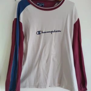 Champion långärmad tröja i vit och vinrött - Snygg långärmad tröja från Champion i vit med vinröda och blå detaljer på ärmarna. Stor Champion-logga tryckt på bröstet. Rund halsringning med vinröd kant. Perfekt för dig som gillar sportig och avslappnad stil.