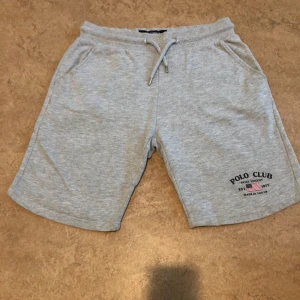 Grå shorts från Polo Club Saint Vincent - Snygga ljusgrå shorts från Polo Club Saint Vincent med snörning i midjan och två sidofickor. Mjukt material i bomull, perfekt för chill dagar. Tryckt logga på ena benet ger en sportig vibe.