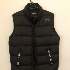 Svart dunväst från WOXO 720° - Svart dunväst från WOXO 720° med hög krage och dragkedja framtill. Västen har två fickor med svarta och vita detaljer på dragkedjorna. Quiltad design ger en schysst look och extra värme. Perfekt att slänga över en hoodie när det är kyligt ute.