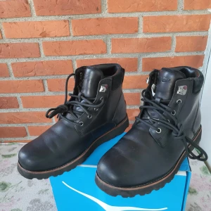 Svarta boots från UGG i skinn - Snygga svarta boots från UGG i skinn med grov sula och snörning. Vattentäta och med UGG-logga bak på hälen. Perfekta för höst och vinter, med robust design och bekväm passform. Klassisk rund tå och detaljer i metall vid snörningen.