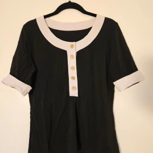 Svart och vit blus med guldknappar från Mexiko - Stilren svart blus med vita detaljer vid halsringning och ärmslut. Inköot i Mexiko. Framsidan har fem dekorativa guldknappar med repmönster. Blusen har korta ärmar och en rak passform. Skulle uppskatta storlek S-M