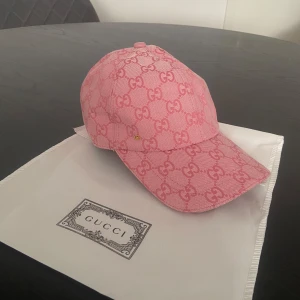 Rosa keps från Gucci med GG-mönster - Snygg rosa keps från Gucci med ikoniskt GG-mönster över hela kepsen. Kepsen är tillverkad i bomull och har justerbar rem baktill. Perfekt accessoar för att lyfta din outfit med en lyxig touch.