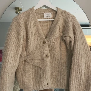 Beige kofta med rosetter från Zara - Supermysig beige stickad kofta från Zara med v-ringning, stora knappar och två snygga rosetter framtill. Koftan har långärmad passform och är perfekt för lager-på-lager-stil. Den fluffiga strukturen ger en trendig och chill vibe.