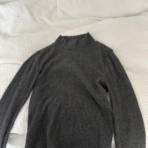 Mörkgrå ribbad polotröja - Mörkgrå ribbad polotröja från Gina Tricot i storlek XS. Tröjan har lång ärm och hög krage, perfekt för lager-på-lager. Tillverkad i mjuk och stretchig mix av polyester, viskos och elastan. Enkel att styla till både jeans och kjol.