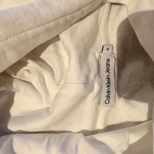 Vit hoodie från Calvin Klein Jeans - Vit hoodie från Calvin Klein Jeans med stor logga i svart och grönt framtill. Klassisk huva och långa ärmar, tillverkad i mjuk bomull. Perfekt för en chill och stilren look.