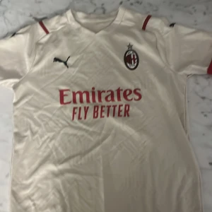 AC Milan vit bortatröja Puma - Säljer en AC Milan bortatröja i vitt från Puma med klubbmärke och Puma-logga på bröstet. Tröjan har röda detaljer på axlarna och Emirates Fly Better-tryck på framsidan. Tillverkad i lätt och ventilerande material, perfekt för fotboll eller supporterliv.