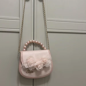 Ljusrosa handväska med pärlhandtag - Supergullig ljusrosa handväska från Elenore med pärlhandtag och dekorativa tygblommor framtill. Väskan har guldfärgad kedja som axelrem och är tillverkad i ett glansigt syntetmaterial. Perfekt accessoar för att lyfta din outfit!