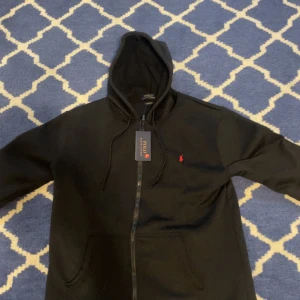 Svart hoodie från Polo Ralph Lauren - Svart hoodie från Polo Ralph Lauren i storlek L. Klassisk design med huva, dragkedja och känguruficka. Broderad röd logga på bröstet och snörning i huvan. Custom fit-modell i mjukt bomullsmaterial, perfekt för chill dagar.