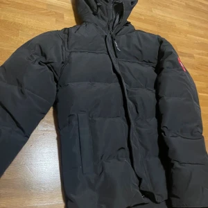 Svart dunjacka från Canada Goose - Säljer en svart dunjacka från Canada Goose med klassisk patch på ärmen. Jackan har hög krage, dragkedja och stora fickor framtill. Perfekt för kalla dagar och riktigt snygg till streetwear-stilen. Pris kan diskuteras vid snabbköp