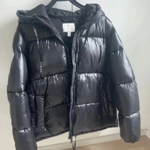 Svart pufferjacka med huva - Säljer en svart pufferjacka med glansig finish och huva. Jackan har dragkedja framtill, långa ärmar och breda, quiltade sektioner som ger en riktigt fet look. Perfekt för kalla dagar när du vill hålla stilen och värmen.