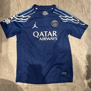 PSG x Jordan blå matchtröja D. Doué - Säljer en blå PSG x Jordan fotbollströja med D. Doué och nummer 14 på ryggen. Tröjan har korta ärmar, vita detaljer och vingar på axlarna. Tillverkad i Dri-Fit material för bästa komfort. Qatar Airways sponsortryck på bröstet.