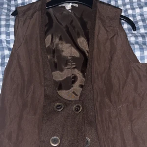 Brun väst från Bandolera med knappar - Snygg brun väst från Bandolera med dubbla rader knappar framtill och justerbart spänne i ryggen. Västen har en klassisk passform och är fodrad med glansigt tyg. Perfekt för att lyfta din outfit med en stilren touch. Storlek 40 men sitter mer som en M