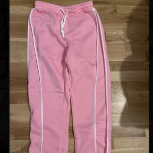 Rosa mjukisbyxor med vita ränder - Säljer ett par ljusrosa mjukisbyxor med vita ränder längs sidorna och vit snörning i midjan. Byxorna har en avslappnad passform och är perfekta för chill dagar. Supermjuka och sköna att ha på sig.