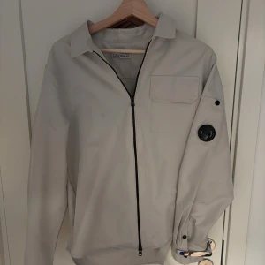 Beige overshirt från C.P. Company - Stilren beige overshirt från C.P. Company med svart dragkedja, bröstficka och den ikoniska linsen på ärmen. Perfekt för lager-på-lager och modern streetstyle. Storlek M i mycket bra skick då den inte kommit jätte mycket till användning. Bara att skriva vid funderingar!!