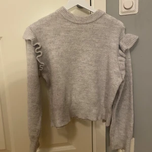 Ljusgrå stickad tröja med volang - Supermjuk ljusgrå stickad tröja från Gina Tricot med volangdetaljer på axlarna. Tröjan har ribbade muddar och en klassisk rund halsringning. Perfekt för dig som gillar en söt och trendig stil.