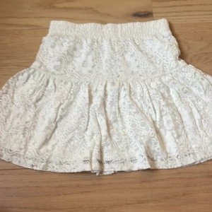 Vit spetskjol med volang, strl 158/164 - Supersöt vit kortkjol med spets och volang från C i storlek 158/164. Kjolen har ett elastiskt midjeband och ett detaljerat spetsmönster med blommor. Perfekt för dig som gillar romantisk och feminin stil.