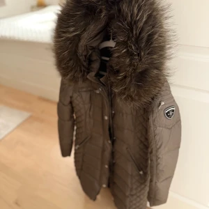 Brun lång dunjacka med fuskpäls - Lång brun dunjacka från Rockandblue med quiltade detaljer och stor huva med fuskpäls. Jackan har dragkedjor på fickorna, tryckknappar framtill och en patch på ärmen. Perfekt för kalla vinterdagar och riktigt snygg passform.