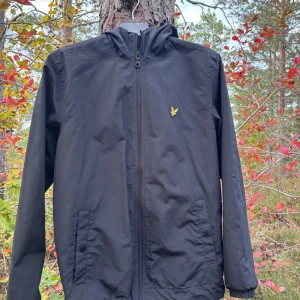 Svart vindjacka från Lyle & Scott S - Svart vindjacka från Lyle & Scott med huva och dragkedja framtill. Jackan har två fickor med knappstängning och den klassiska gula loggan på bröstet. Perfekt för blåsiga dagar och enkel att matcha med allt. Materialet är lätt och känns smidigt.