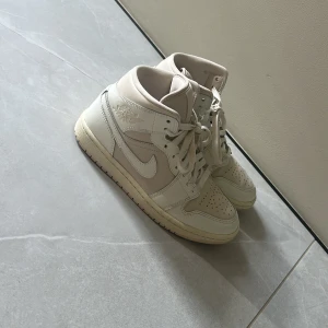 Nike Air Jordan 1 Mid Beige 38.5 - Säljer ett par Nike Air Jordan 1 Mid i en snygg beige och vit färg. Skorna har klassisk snörning, perforerad tå och ikoniska detaljer som Jordan-loggan på sidan. Tillverkade i skinn med en mjuk vadderad insida för extra komfort. Perfekta för dig som gillar streetstyle.
