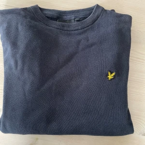 Mörkgrå stickad tröja från Lyle & Scott - Mörkgrå stickad tröja från Lyle & Scott med klassisk rund hals och gul broderad logga på bröstet. Tröjan har ribbade muddar vid ärmslut och nederkant, perfekt för dig som gillar stilren och enkel design.