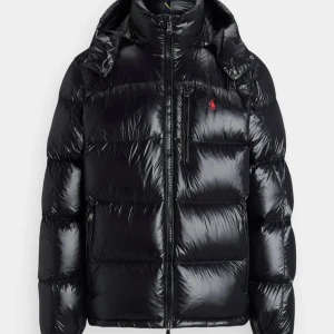 Svart pufferjacka från Polo Ralph Lauren - Snygg svart pufferjacka från Polo Ralph Lauren med glansig finish och röd logga på bröstet. Jackan har hög krage, huva och dragkedja framtill samt praktiska fickor med dragkedja. Perfekt för kalla dagar när du vill hålla stilen.