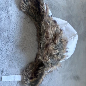 Vit pälsfodrad vinterhatt KappAhl - Säljer en vit vinterhatt från KappAhl med mjukt fuskpälsfoder i bruna och grå toner. Hatten har öronlappar, knappar och ett band under hakan. Perfekt för kalla dagar när du vill hålla dig varm och ändå se cool ut.