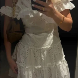 Vit kort klänning med volang - Superfin vit kortklänning i Positano-stil med breda volanger vid axlarna och offshoulder-design. Klänningen har broderade detaljer och är tillverkad i bomull, vilket gör den luftig och skön. Perfekt för sommarens alla tillfällen när du vill känna dig extra söt.