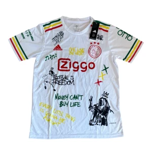 Ajax x Bob Marley vit fotbollströja - Ajax x Bob Marley limited edition fotbollströja i vitt med färgglada ränder på ärmarna. Tryck med Bob Marley, graffiti-inspirerade texter och Ajax-logga på bröstet. Materialet är lätt och andas, perfekt för fotboll eller streetwear. Ziggo sponsortryck på framsidan.