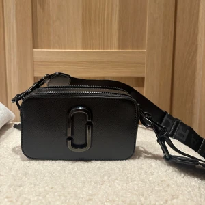 Svart axelväska från Marc Jacobs - Snygg svart axelväska från Marc Jacobs med stilren logga framtill. Väskan har en bred axelrem med logotyp, dubbla dragkedjor och är tillverkad i strukturerat skinn. Perfekt för att bära det viktigaste med stil. Köpt för 4500kr