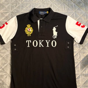 Svart och vit polo från Ralph Lauren - Polo från Ralph Lauren i svart och vitt med röda detaljer och broderad Tokyo-text framtill. Klassisk krage, korta ärmar och stora siffran 5 på ryggen och ärmarna. Slim fit-modell i bomull, perfekt för dig som gillar sportig och stilren look.