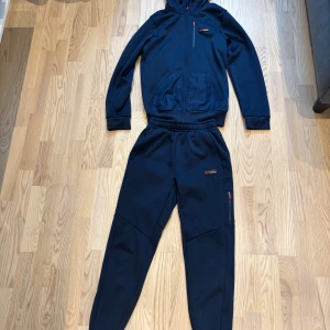 Mörkblå mjukisbyxor från McKenzie - Säljer ett par mörkblå mjukisbyxor från McKenzie med elastisk midja och dragsko. Byxorna har en sportig look med fickor på sidorna och diskret logga på benet. Perfekta för chill eller träning, tillverkade i mjukt syntetmaterial.