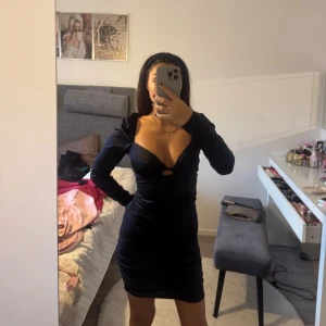 Svart figurnära kort klänning - Säljer en svart, figurnära kort klänning med lång ärm och djup v-ringning. Klänningen har en snygg cut-out detalj framtill och är gjord i ett glansigt, stretchigt material som ger en lyxig känsla. Perfekt för dig som vill sticka ut med en stilren look.