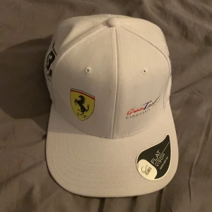 Vit Ferrari keps Gran Turismo  - Snygg vit keps med platt skärm, dekorerad med Ferrari-logga, Gran Turismo Campionato Italiano-tryck och flera coola patches på sidorna. Justerbar baktill och tillverkad i bomull. Perfekt för motorsportfans som vill sticka ut. Limited edition. Som inte finns att köpa!